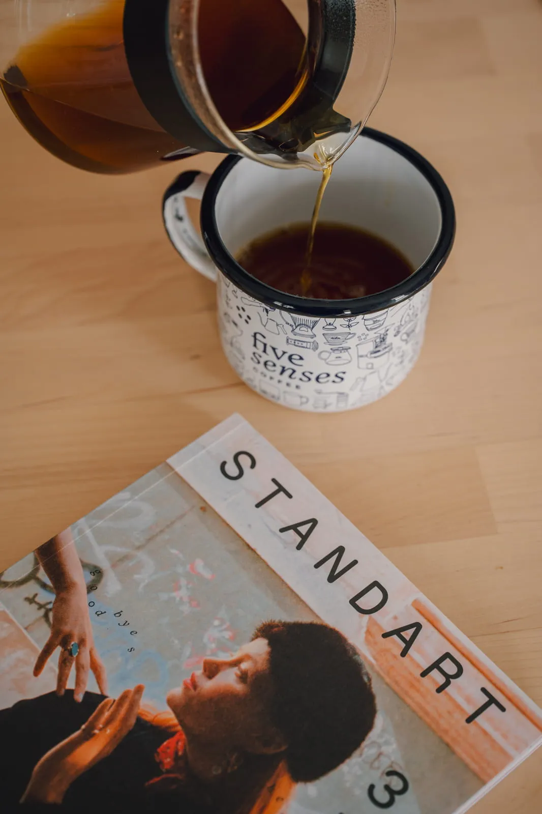 Coffee poured from a glass carafe into a Five Senses enamel mug, with Standart magazine in the foreground