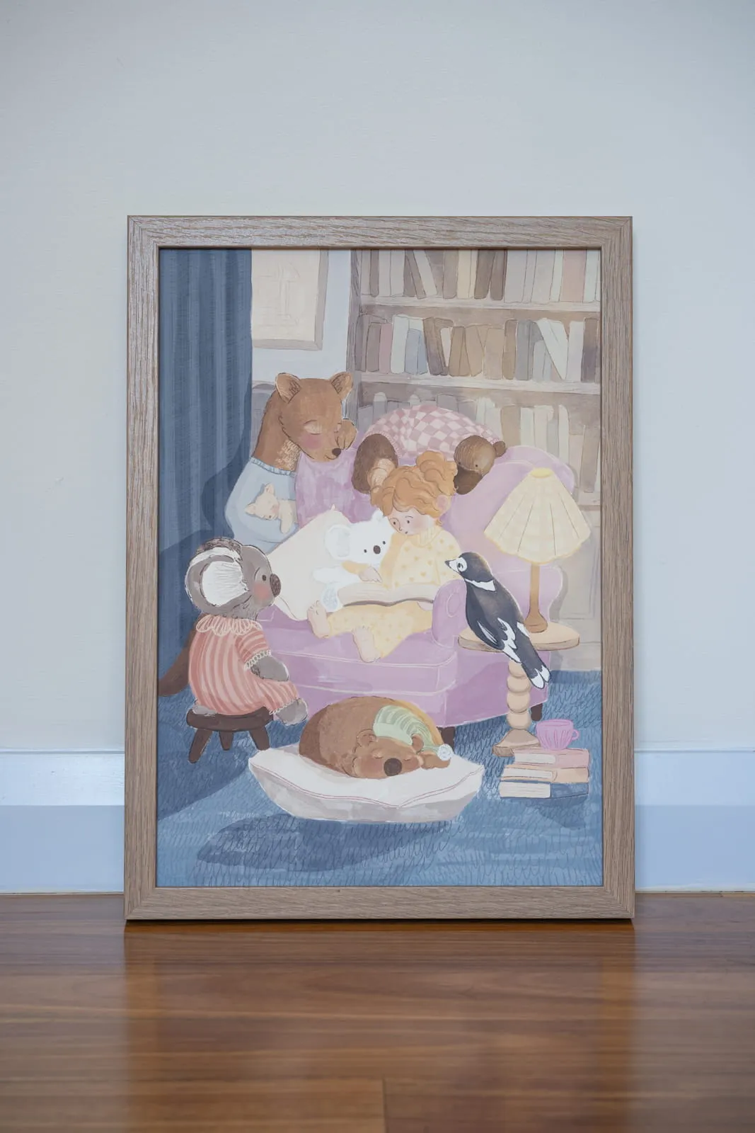 Framed illustrated artwork of animals gathered around a bookshelf in a cosy reading scene, leaning against a white wall