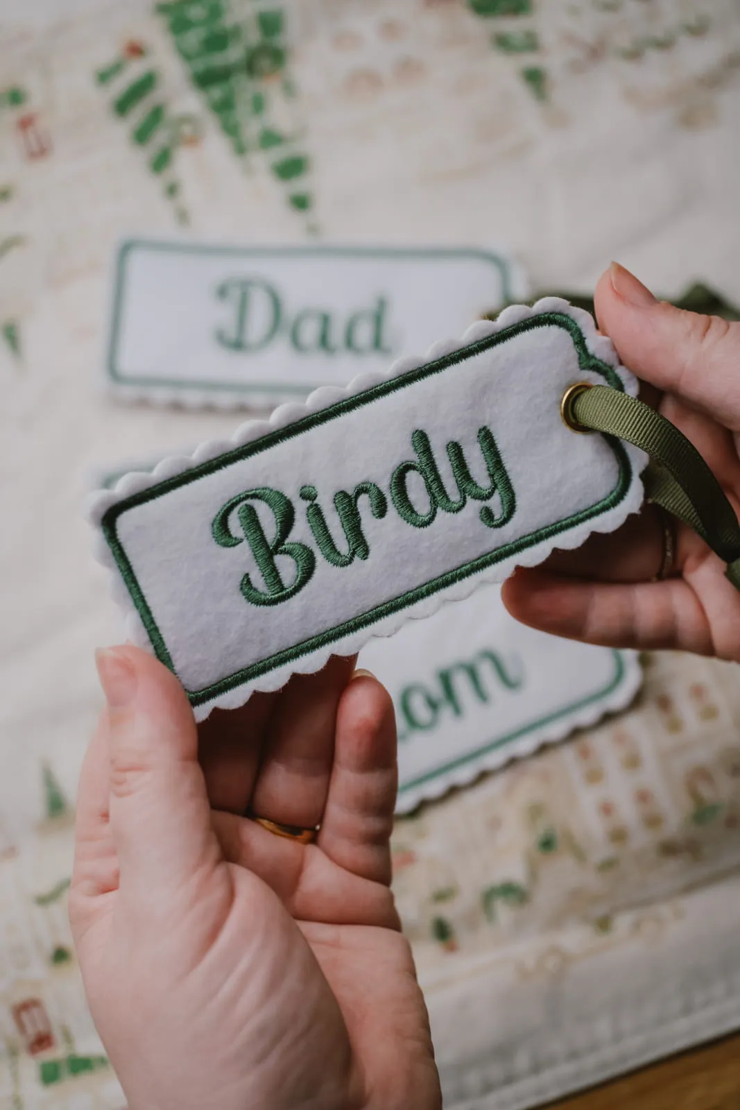 Hands holding an embroidered felt gift tag reading Birdy, with more personalised tags visible in the background