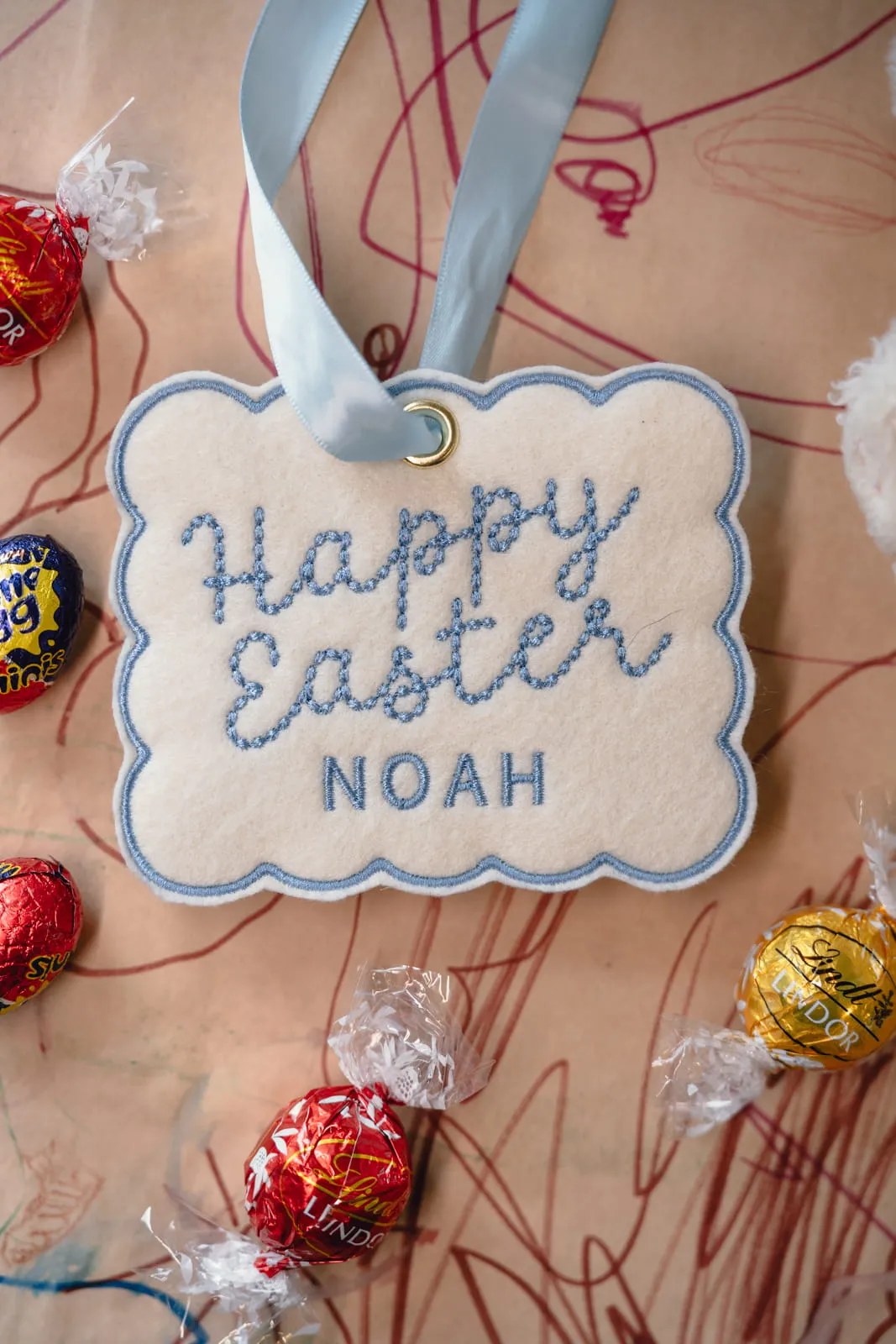 Happy Easter Noah embroidered felt tag with blue scalloped border lying on a child's crayon-drawn paper