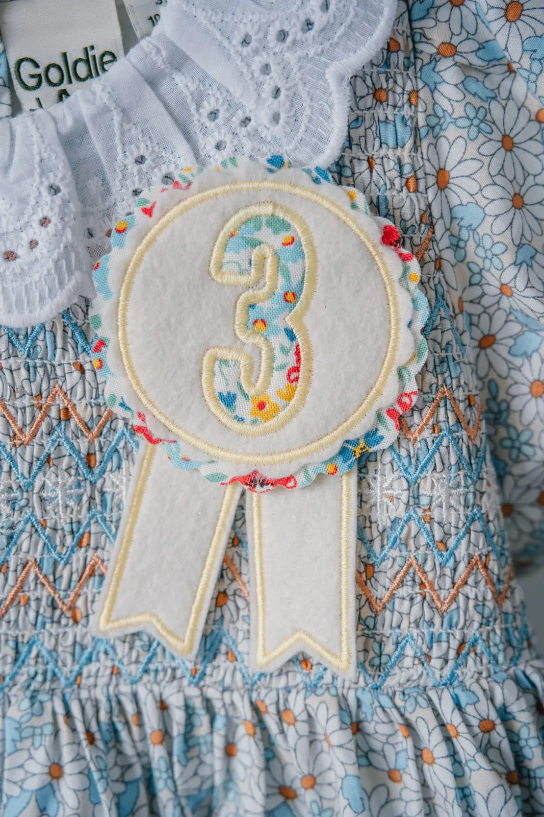 Child wearing a floral dress with an embroidered number 3 birthday rosette badge pinned to it