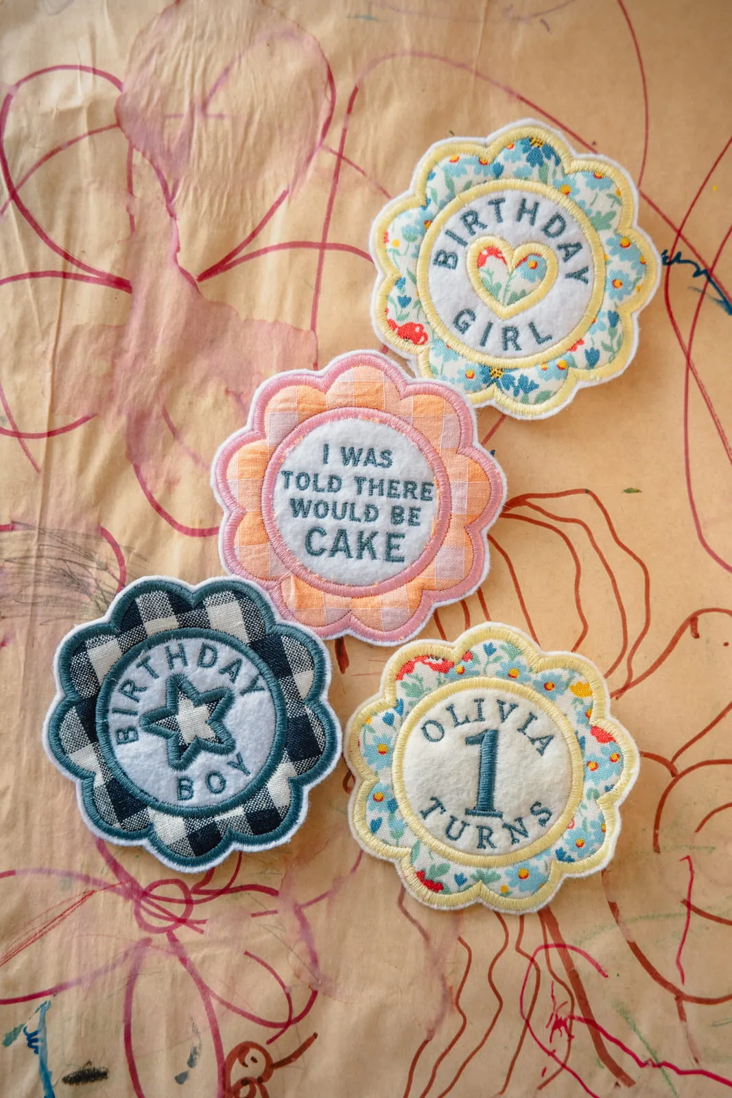 Flat lay of four scallop-edge embroidered birthday badges including Birthday Girl, I Was Told There Would Be Cake, and Olivia Turns 1