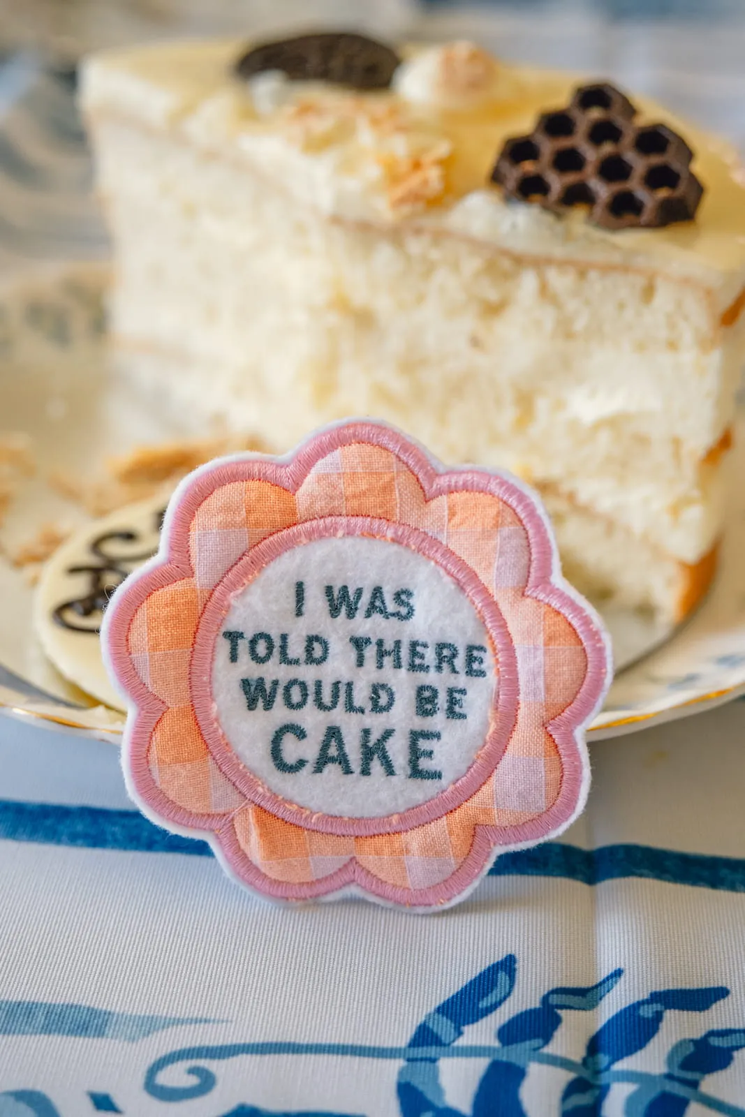 I Was Told There Would Be Cake embroidered patch propped against a slice of white cake on a blue floral plate