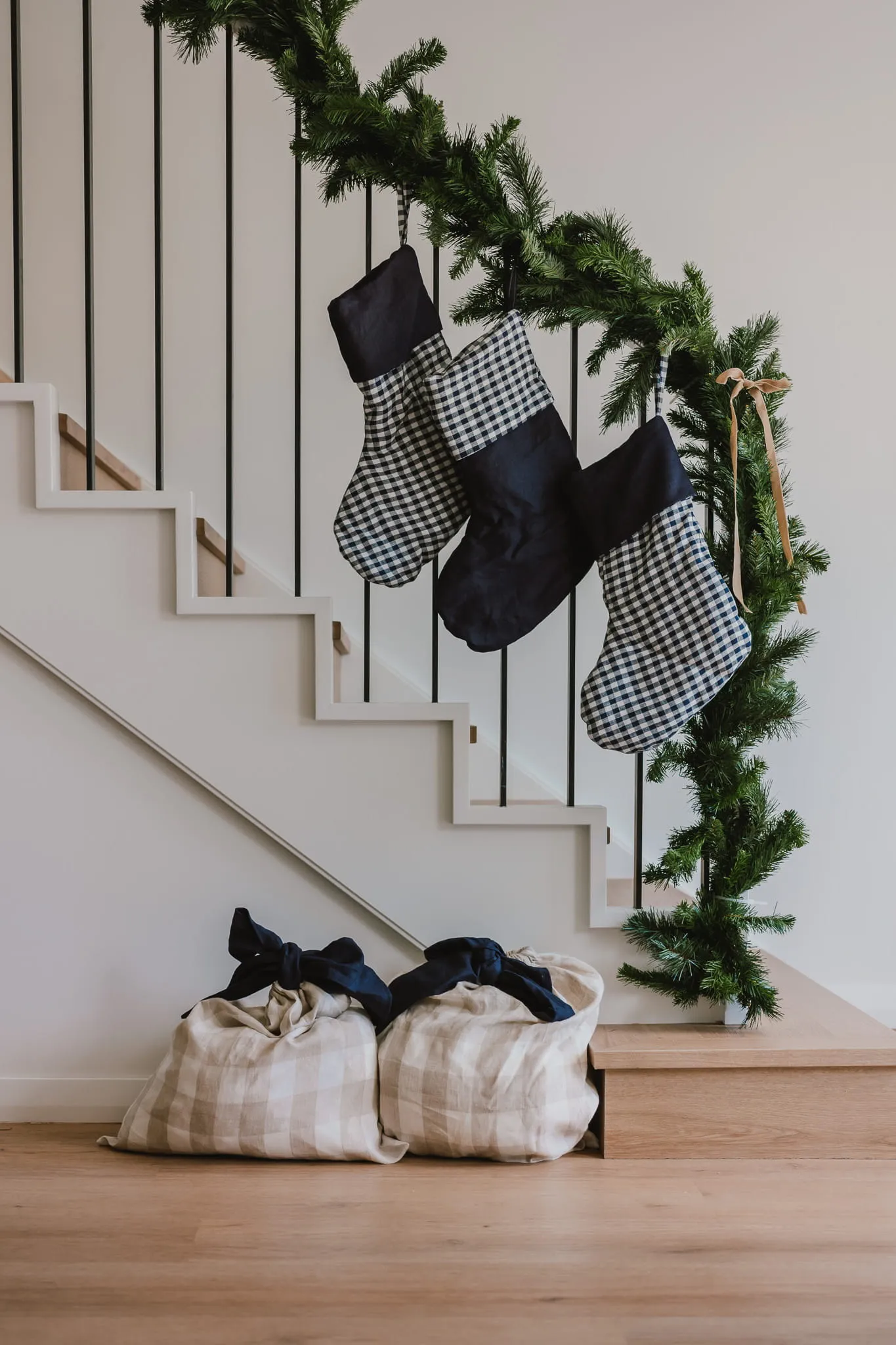 Three gingham and navy Christmas stockings hanging from a stair banister with pine garland, with plaid Santa sacks below